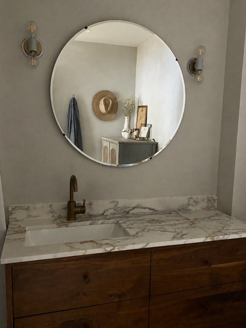 A round beveled mirror flanked by brass sconces above a marble vanity with a brass faucet and walnut cabinetry.