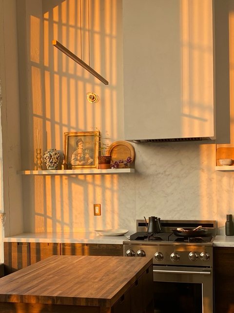 Golden-hour light through a window screen striping the range hood and marble backsplash, with a framed portrait and ceramics on the open shelf.