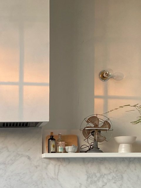 Window-screen shadows on the range hood and wall beside a marble shelf styled with a vintage fan, glass bottles and a small ceramic bowl.