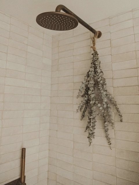 An antique-bronze rain shower head against zellige subway tile, with a bundle of dried eucalyptus hanging from the arm.