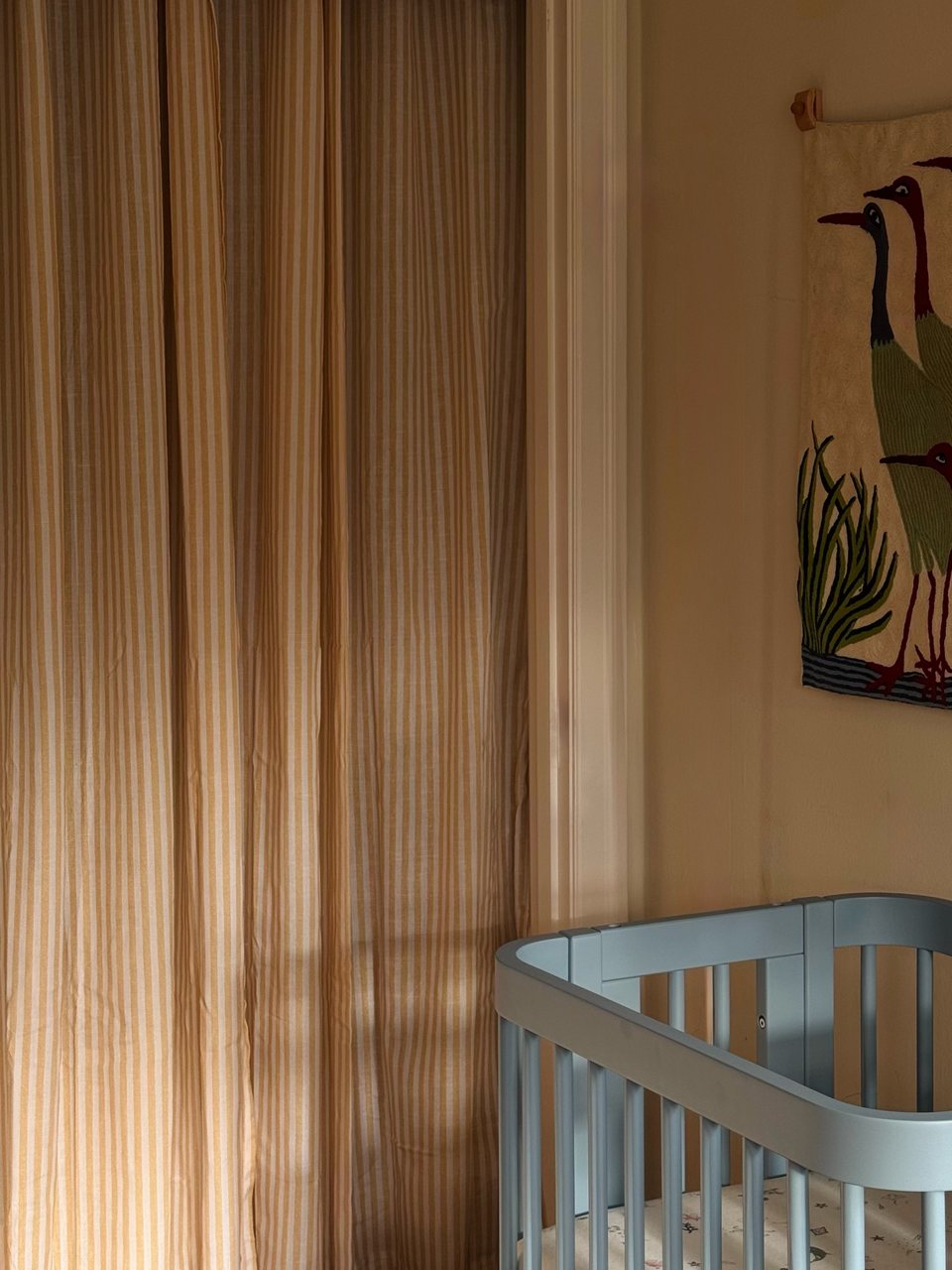 Striped mustard and cream linen curtains beside a door frame, with a blue wooden crib and an embroidered heron wall hanging in a warm peach nursery.